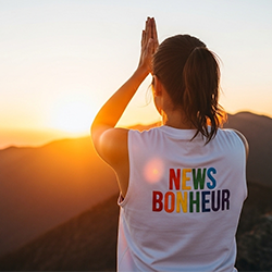 femme saluant un coucher de soleil sur des montagnes avec l'inscription "News Bonheur" brodée en lettre de couleur sur son t-shirt